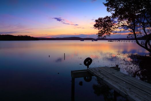 Sunset reflections at sundown over Kincumber broadwater.  A little jetty in the foreground, boats and a larger jetty in the background.