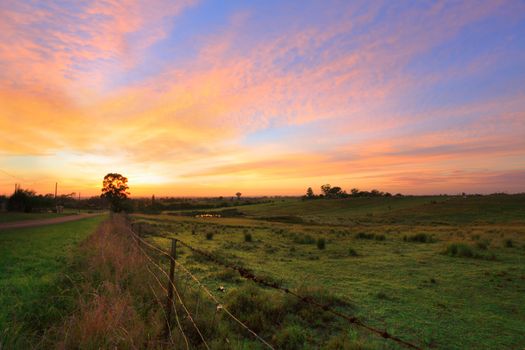 Sunrise throws a warm light over the countryside hills, paddocks and farm sheds.   Orchard Hills, Australia