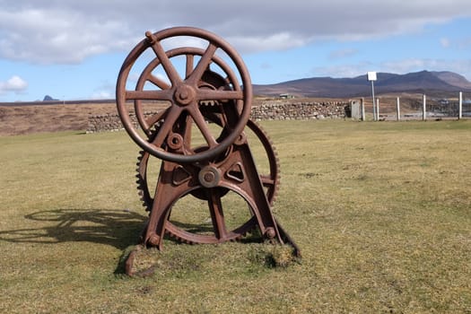 A vintage, antique hand winch, rusty metal, standing on a patch of short green grass.