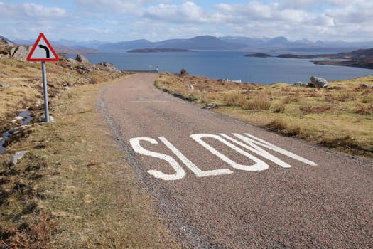 A narrow road with the word 'SLOW' painted on the floor and a triangular sign indicating a bend ahead with the sea and islands in the distance.