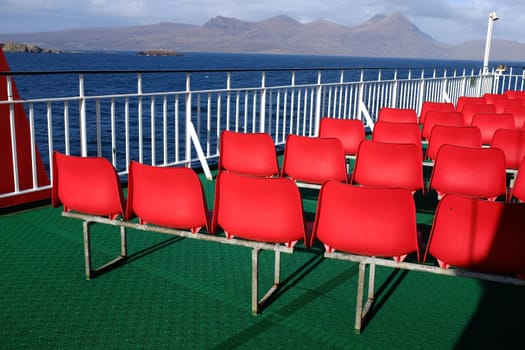 Rows of red plastic seating on a green rubber floor with white railings and a view of the sea and mountains in the distance.