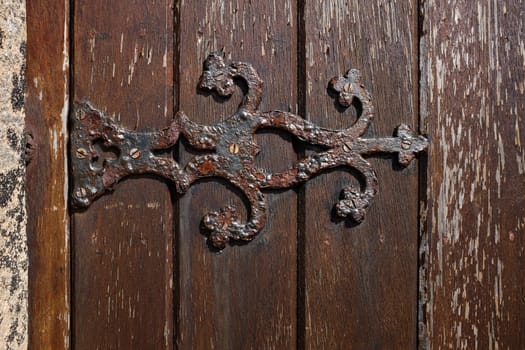 An ornate metal hinge with rust and flaking paint attached to a stained wooden door.