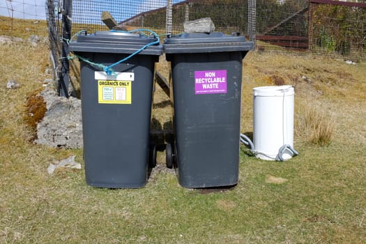 A pair of waste bins with colour coded signs, yellow with the words 'ORGANICS ONLY' and purple with 'NON RECYCLABLE WASTE' on grass infront of a fence.