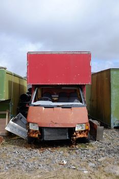 A red van broken and wrecked with a broken windscreen between two green containers against a grey cloudy sky.