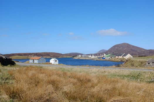 Grasses lead to tin huts overlooking a small loch with the village of Leverburgh, Isle of Harris, Scotland, UK.