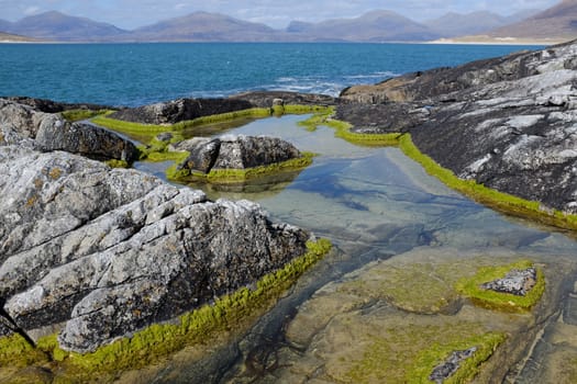 A rock pool with a rim of green algae leading to the sea and mountains beyond.