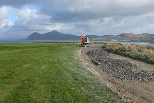 A cliff top track with new hardcore spread over mud, a mechanical digger is parked on grass next to the track with a view over the sea to mountains in the distance.
