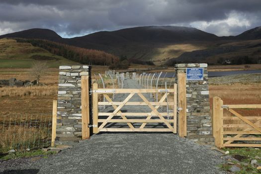 A wooden gate leads to a causeway with a hardcore path flanked by metal fencing with cloud covered mountains in the distance.