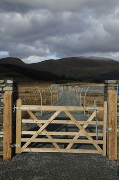 A wooden gate leads to a causeway with a hardcore path flanked by metal fencing with cloud covered mountains in the distance.