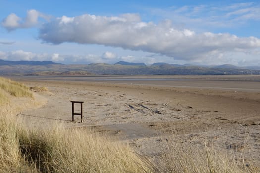 Sand dune grasses lead to a large deserted beach with empty sign post, a mountain range can be seen in the distance.