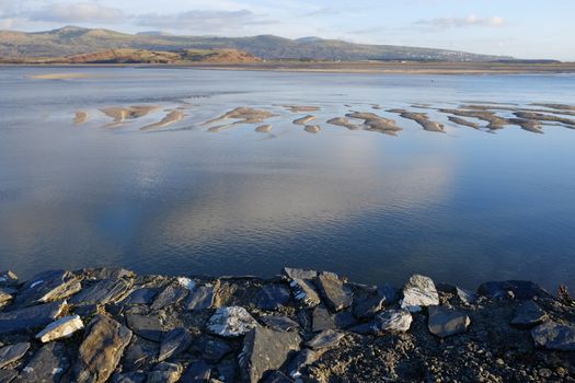 A rocky view point looks out across an estuary with sand bars being covered by the incoming tide. The town of Harlech can be seen in the distance with the Rhinnog mountain range, Snowdonia national park, Gwynedd, Wales, UK, across the horizon.