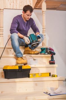 a young carpenter sitting on step of wooden ladder holding and looking on woodworking device