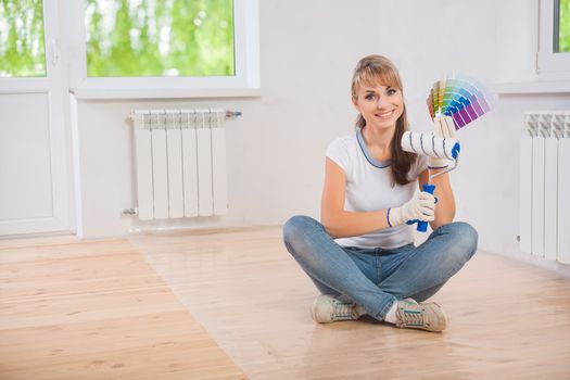 beautiful smiling female worker sitting on wooden floor and showing paintroller and color palette