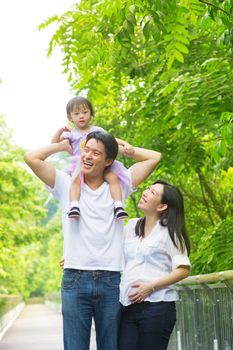 Happy Asian family outdoor. Father piggyback his daughter walking in garden park with pregnant wife. Healthy lifestyle. 