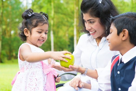 Happy Indian family. Asian girl sharing an green apple at outdoor with mother and sibling.