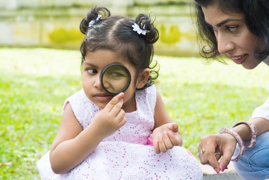 Daughter at outdoor green park with mother. Cute Indian girl peeking through magnifying glass with parent.