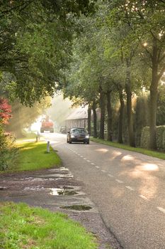 Country road with traffic, farm, trees and sunbeams at sunrise 