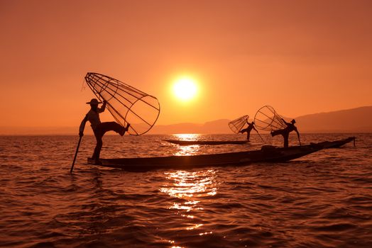 Silhouette of traditional fishermans in wooden boat using a coop-like trap with net to catch fish in Inle lake, Myanmar