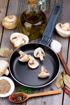 Raw Edible Champignon Mushrooms in Frying Pan, Spices in Wooden Spoons, Ingredients and Olive Oil closeup on Rustic Wooden background