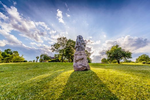 Idyllic landscape of the park with stone sculpture in the centre, shot against sun, visible green trees, green grass with small white flowers, in background sunrays, blue sky and sharp clouds. Photo taken in public place.