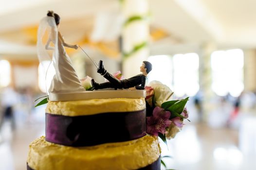 Fragment of yellow and purple wedding cake with funny decoration symbolizing groom pulled by leg on a leash by bride, wedding hall in background.