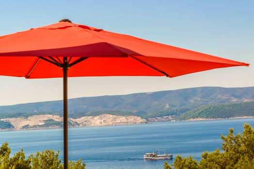 Red terrace, garden umbrella against blue sea and cloudless sky, visible coastline, ship in distance.