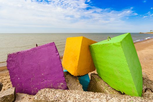 Three different colours - purple, yellow and green concrete breakwater blocks on the beach, in the backgroundblue cloudless sky, yellow sand and coastline.
