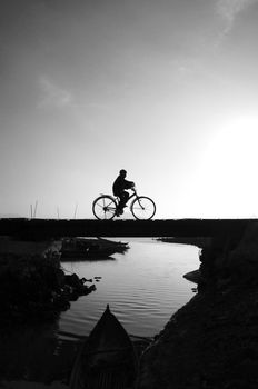 Silhouette of lonely young boy riding bicycle on small bridge cross the river at morning, he ride bike leisure,  this make sad, calm scene at countryside 