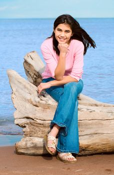 Beautiful biracial teen girl sitting on fallen log by lake shore in summer