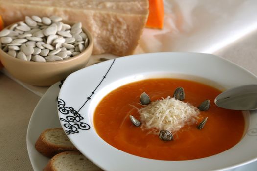 A plate of pumpkin cream soup with pumpkin seeds,two pieces of bread,italian parmesan cheese and a spoon on the plate