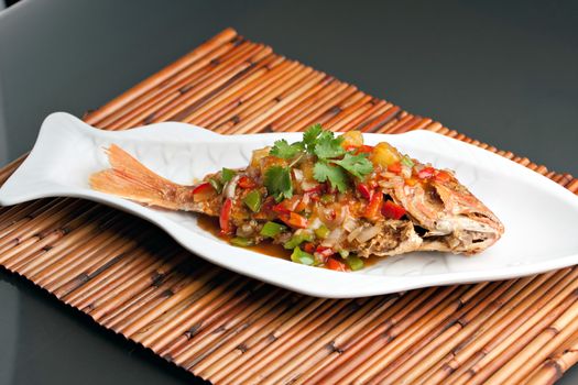 Freshly prepared Thai style whole fish red snapper dinner with tamarind sauce on a white fish shaped plate.