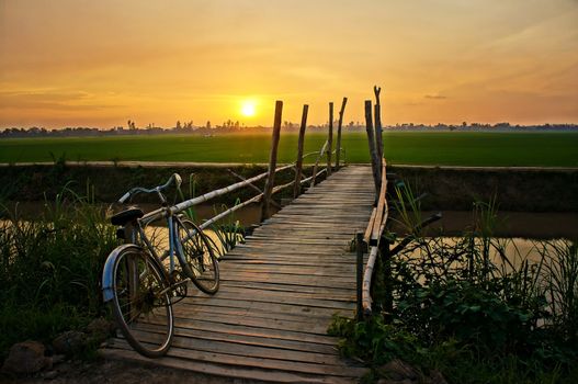 Beautiful landscape of nature with lovely orange sunset, bicycle put up on wooden fence, small wooden bridge across a river and in the distance is green rice field 