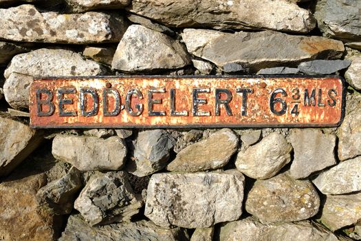 An antique road sign damaged and rusty fixed to a stone wall reading ' BEDDGELERT 6 and 3/4 MLS' in flaked black paint.