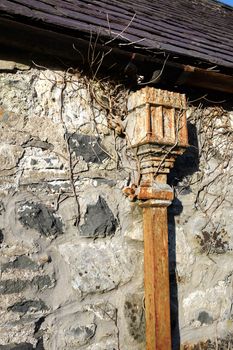 A slate roof slopes down to a damaged cast iron gutter with a decorative water catcher in cast iron attached to a square down pipe and bracket fixed to a traditional lime mortar wall.