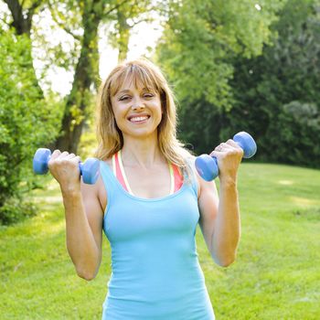 Portrait of female fitness instructor exercising with small weights in green park