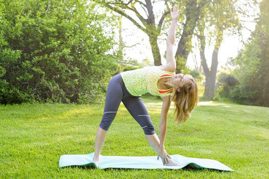 Female fitness instructor doing yoga extended triangle pose outdoors in green park