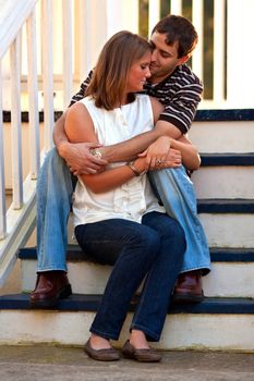 A young couple in love share a romantic embrace while sitting on steps of gazebo. 