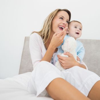 Cheerful mother and baby boy with stuffed toy looking away in bed