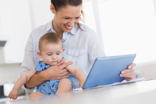 Smiling mother calculating finances on digital tablet while holding baby boy at kitchen counter