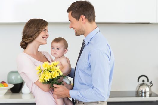 Father giving mother holding baby a bunch of yellow flowers at home in the kitchen