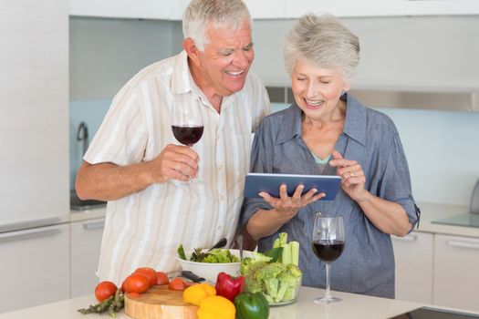 Smiling senior couple preparing a salad and using tablet pc at home in the kitchen