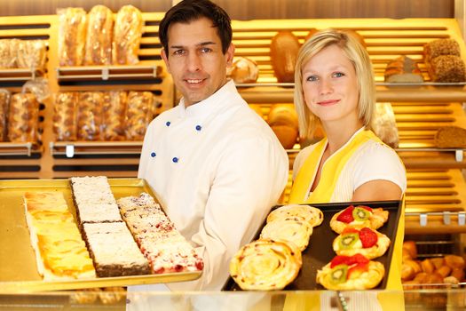 Bakery shopkeeper and baker present different types of pastry in shop