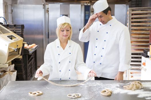 Apprentice in bakery or bakehouse trying to form a pretzel out of dough without success. Her instructor watches.