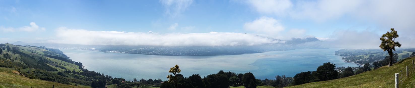 Broad panoramic landscape of the Otago Peninsula and Bay with the city of Dunedin in the distance and clouds hanging over the mountains