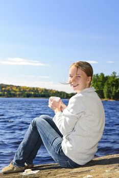 Young woman sitting with beverage on rock relaxing by beautiful lake in Algonquin Park, Canada