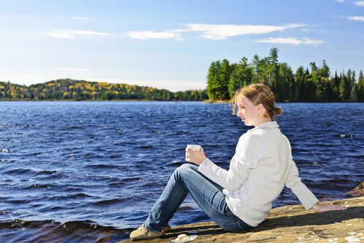 Young woman sitting with beverage on rock relaxing by beautiful lake in Algonquin Park, Canada.