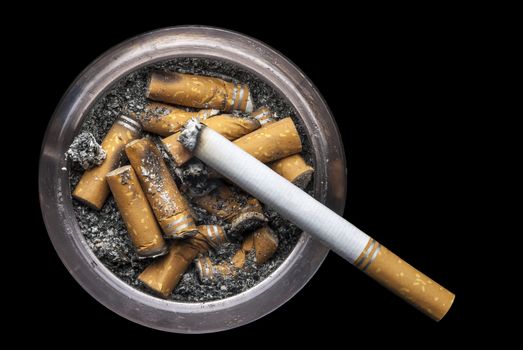 Image of a grungy ashtray full of ash and cigarette butts with one lit cigarette on a black background. Space for copy.