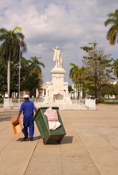 CIENFUEGOS, CUBA - FEBRUARY 25, 2014: A garbage man drags green bin through Plaza Jose Marti in Cienfuegos, Cuba on February 25, 2014