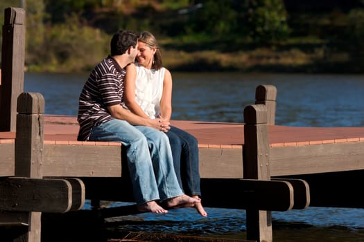 A young couple in love share an intimate moment while sitting barefoot on a dock over a lake.
