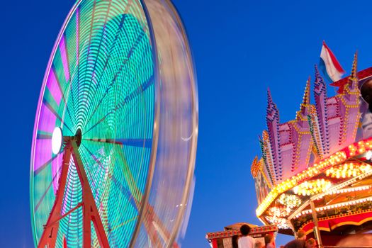 The movement and colorful lights of a carnival ferris wheel motion blur against a twilight sky.  